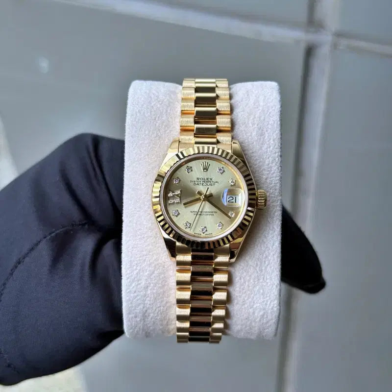 Gold watch on a white cloth held by a gloved hand against a gray tiled wall.