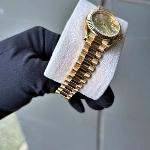 Gold watch on a white cushion with a black glove holding it, on a light gray background